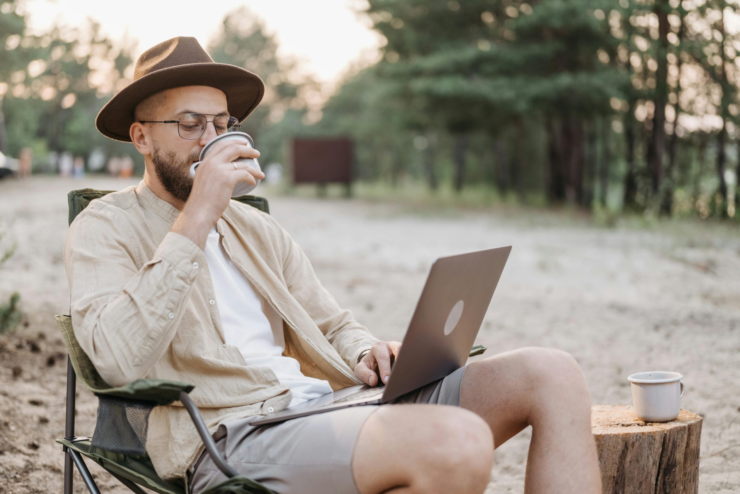 A man sips coffee as he works on his laptop in an outdoor setting.