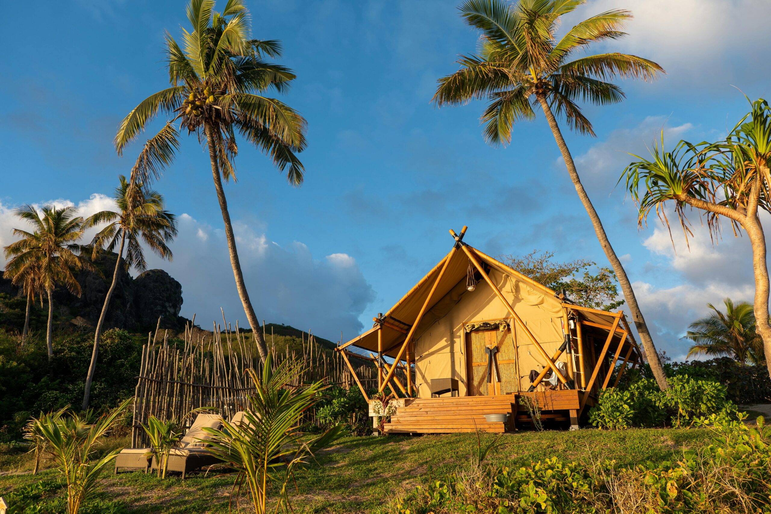 GSA251-Glamping structures in Palm Trees A scenic glamping tent set among palm trees under a blue sky. In the bottom left corner is the Glamping Americas Podcast logo with a photo of host Sarah Riley and a red glamping cabin.
