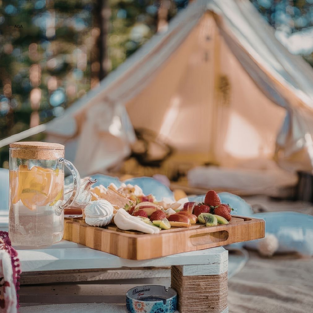 Wooden tray with fresh fruit, pastries and tea set on a rustic table in front of a softly lit glamping tent in a forest setting.