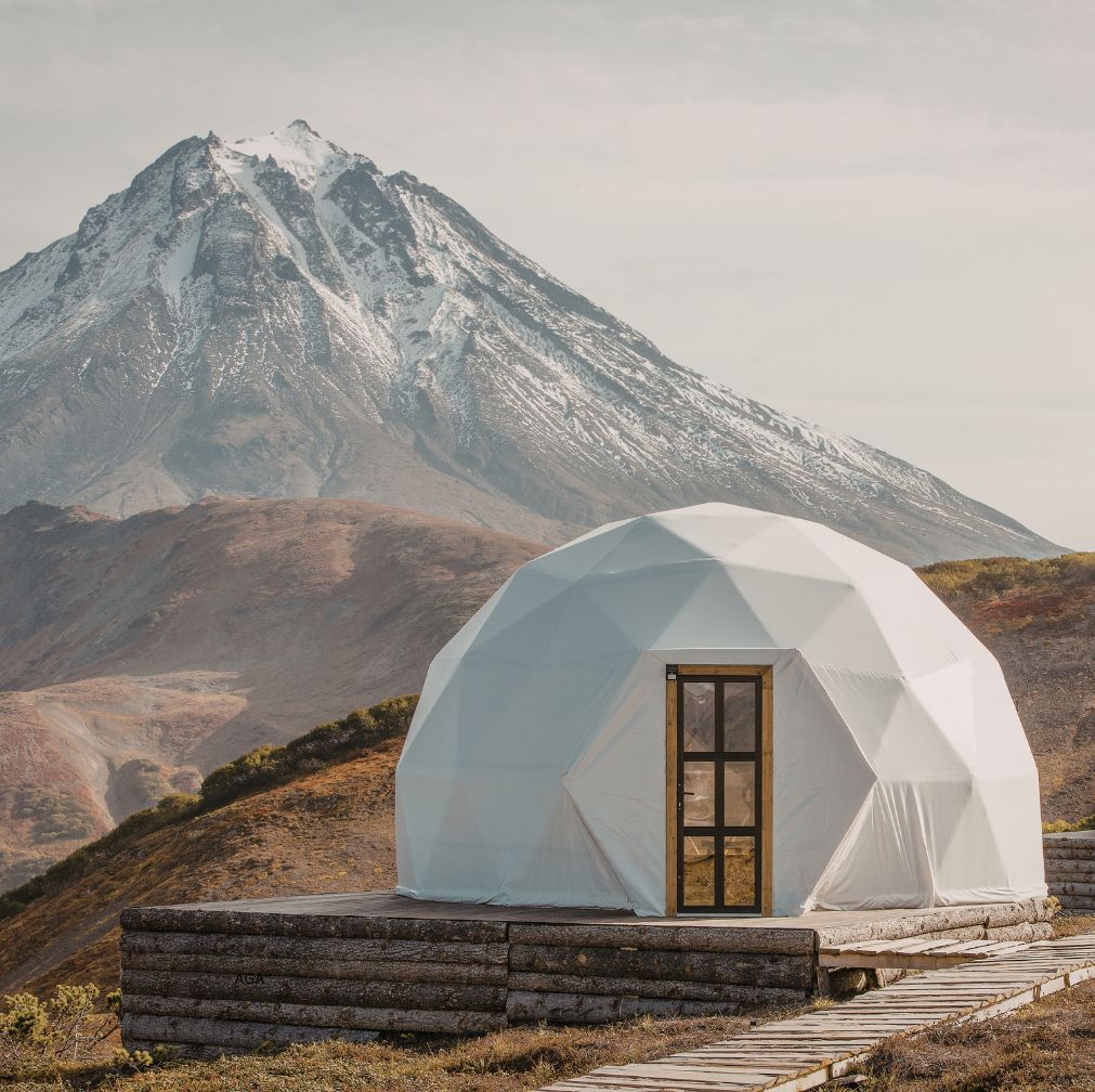 White geodesic dome glamping tent on a raised platform with a mountain landscape and snow-capped peak in the background.
