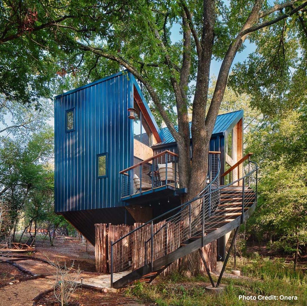 Modern elevated treehouse cabin with dark metal siding and wood accents, built around a large tree, featuring a small deck and staircase, surrounded by lush forest at Onera.