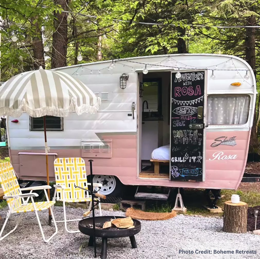 Vintage pink-and-white camper trailer set up in a wooded campsite at Boheme Retreats, with a striped umbrella, two retro lawn chairs, and a small fire pit with logs in front. The camper door is open, revealing a cozy interior, and a chalkboard on the door reads “Adventure with Rosa” with camping-themed doodles.