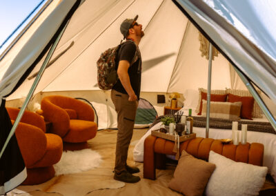 Person standing inside a furnished glamping tent, looking up at the structure. The space features a styled bed with pillows, a tray with wine and decor, and cozy orange lounge chairs, showcasing a comfortable, upscale camping interior.