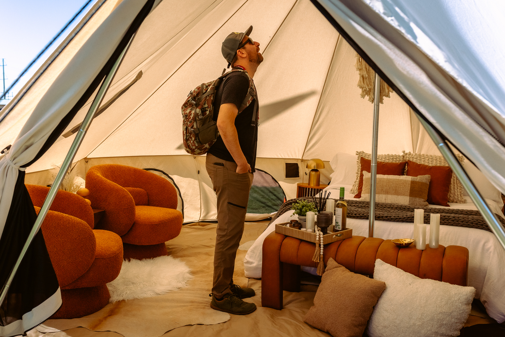 Person standing inside a furnished glamping tent, looking up at the structure. The space features a styled bed with pillows, a tray with wine and decor, and cozy orange lounge chairs, showcasing a comfortable, upscale camping interior.