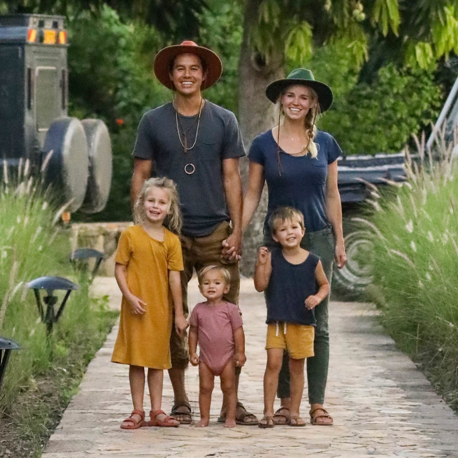 A smiling Bucket List Family—Garret and Jessica Gee and three young children—stand together on a garden pathway, holding hands and wearing casual clothes and wide-brim hats, surrounded by greenery.
