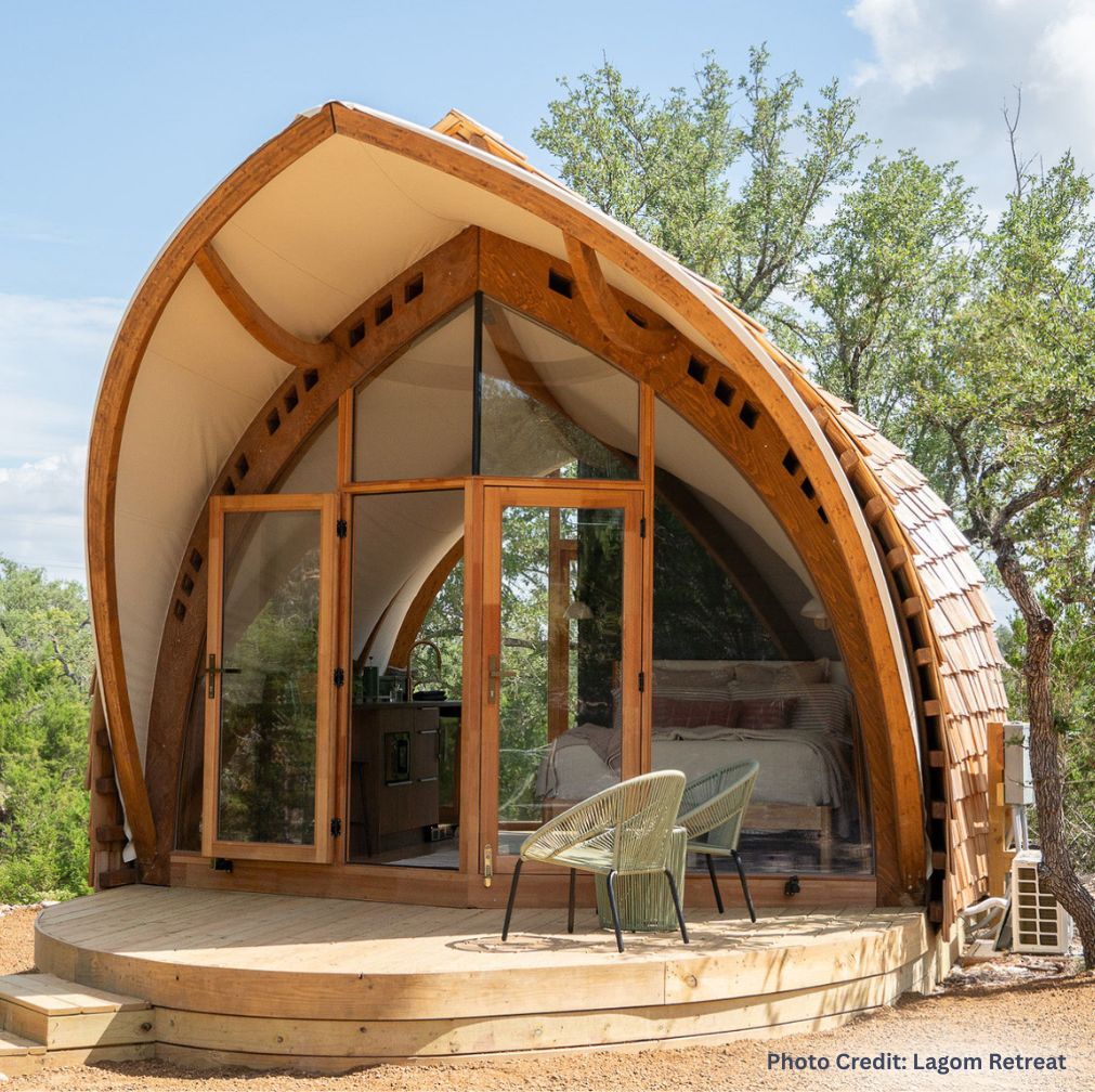 Curved wooden glamping cabin with a glass front, showing a cozy bed and small kitchenette inside, set on a raised deck with two chairs and surrounded by trees at Lagome Retreat.