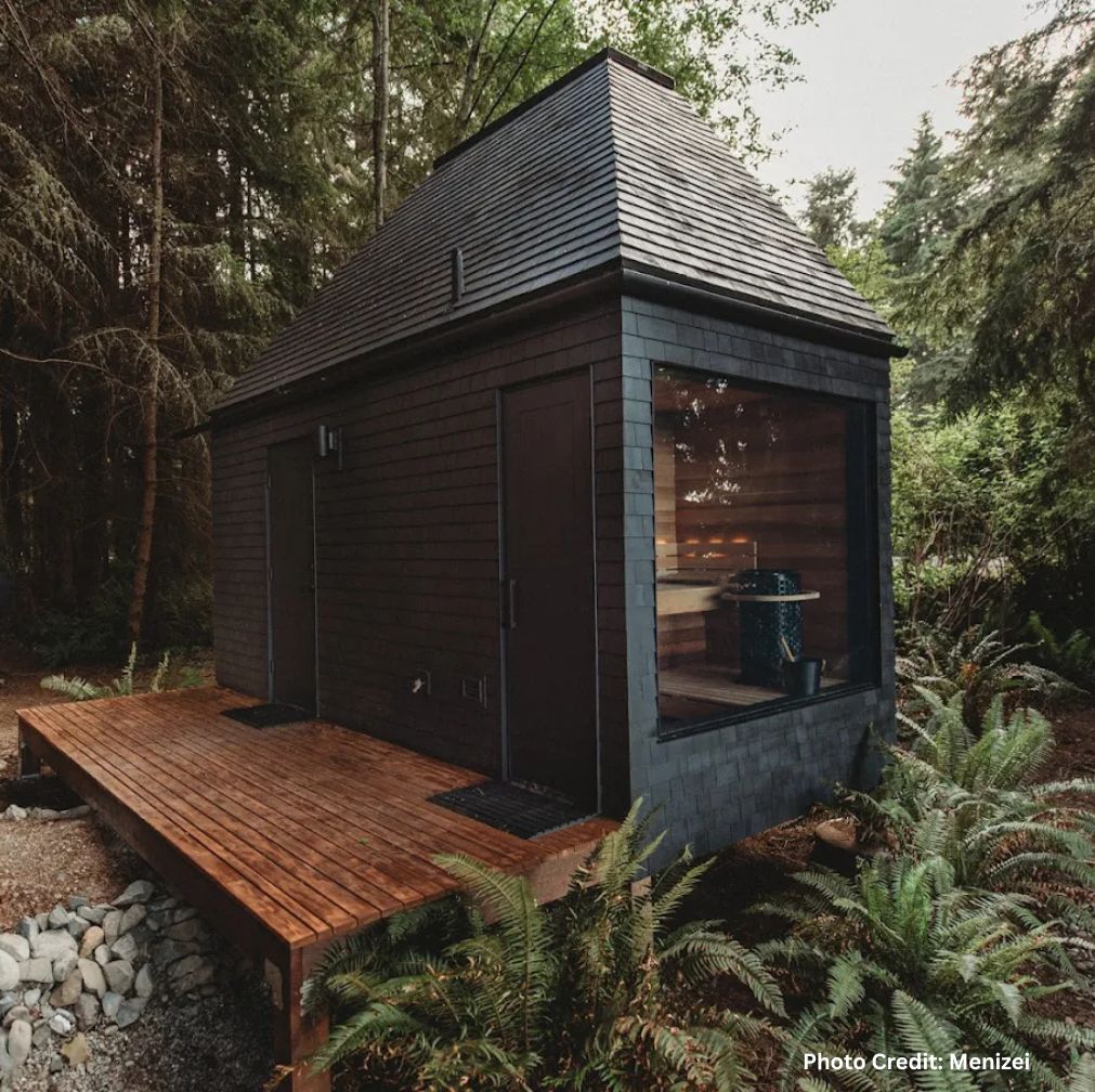 Modern dark wood cabin at Menizei with a steep roof and large window revealing a sauna interior, set on a small deck surrounded by lush forest and ferns.