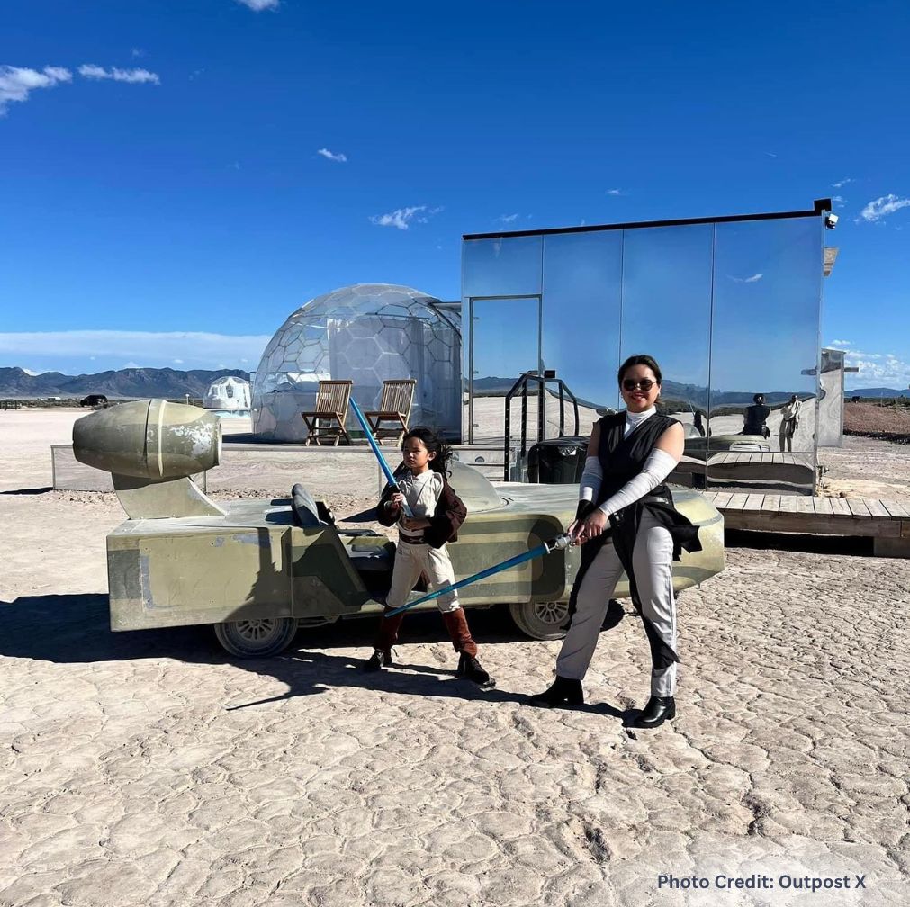 Adult and child posing with toy lightsabers beside a futuristic, speeder-style vehicle in a desert landscape, with mirrored cabins and a geodesic dome in the background. A familiar scene at Outpost X.