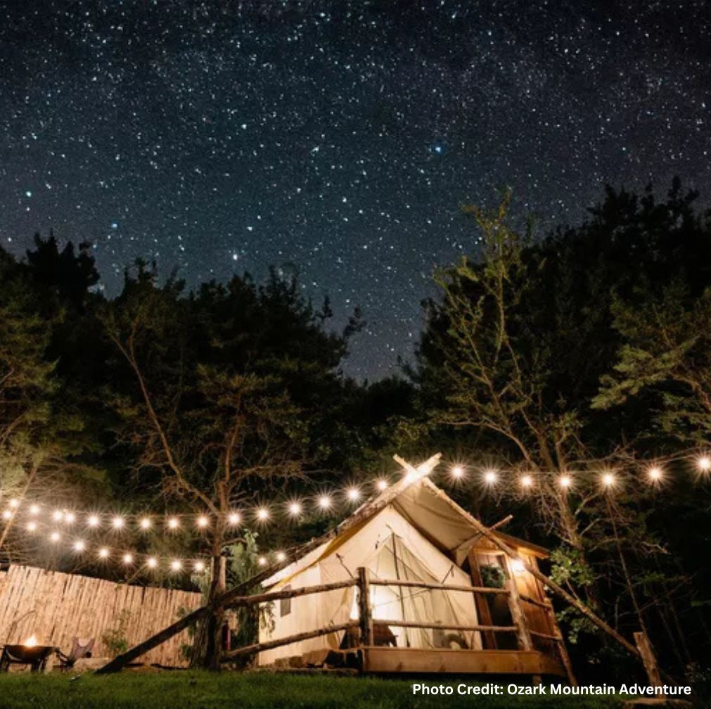 Glamping tent illuminated by string lights in a forest clearing at night, with a fire pit and seating nearby under a sky full of stars at Ozark Mountain Adventure.