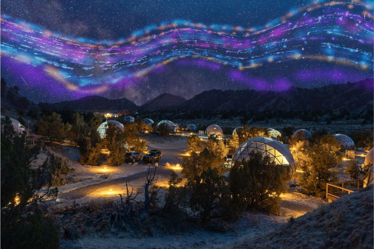 Glamping domes glowing at night in a desert landscape with warm pathway lights, set beneath a vivid starry sky with colorful, stylized light patterns overhead