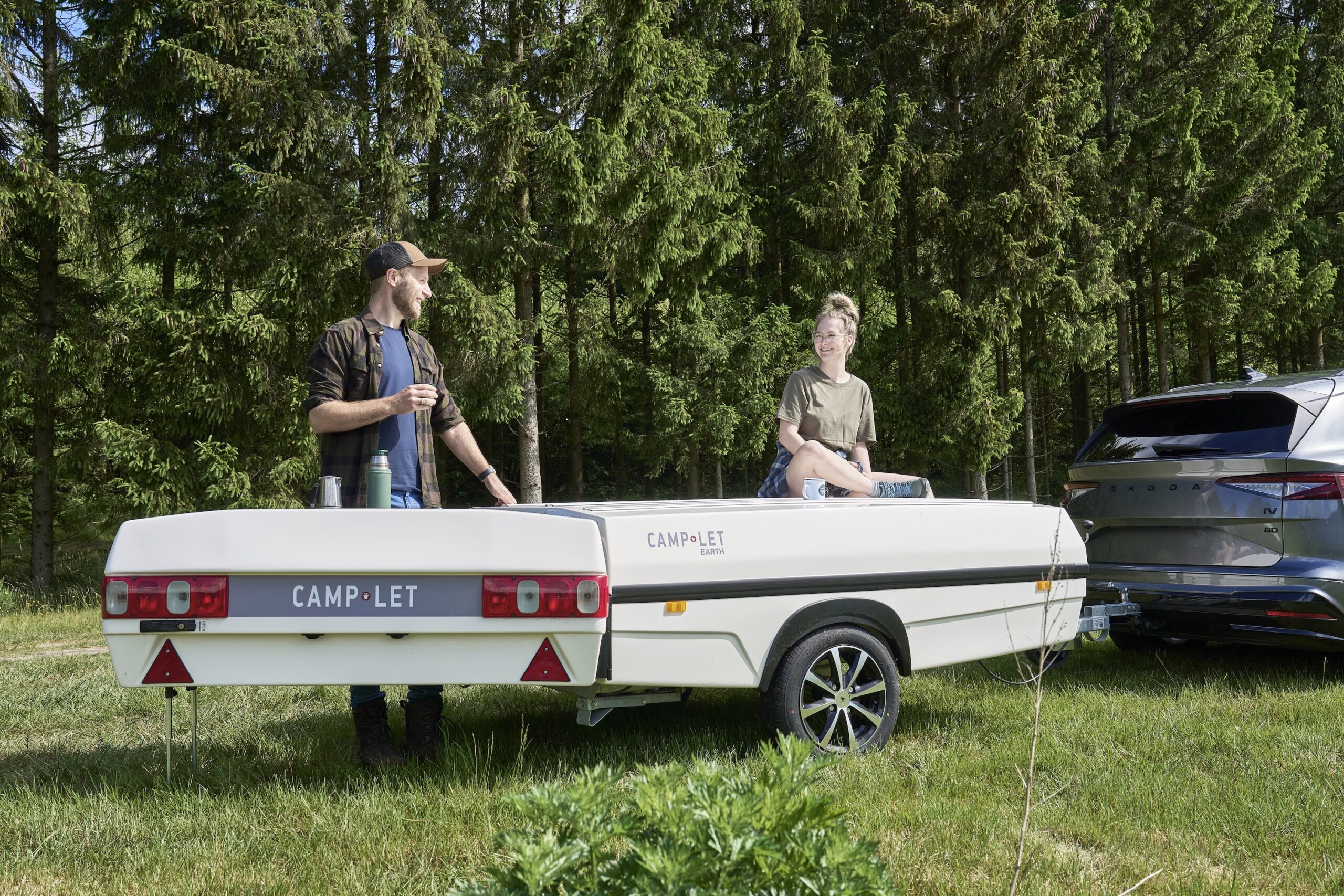 Two people stand and sit on a closed Camp-let tent trailer hitched to a car in a grassy forest clearing, chatting casually with drinks nearby.