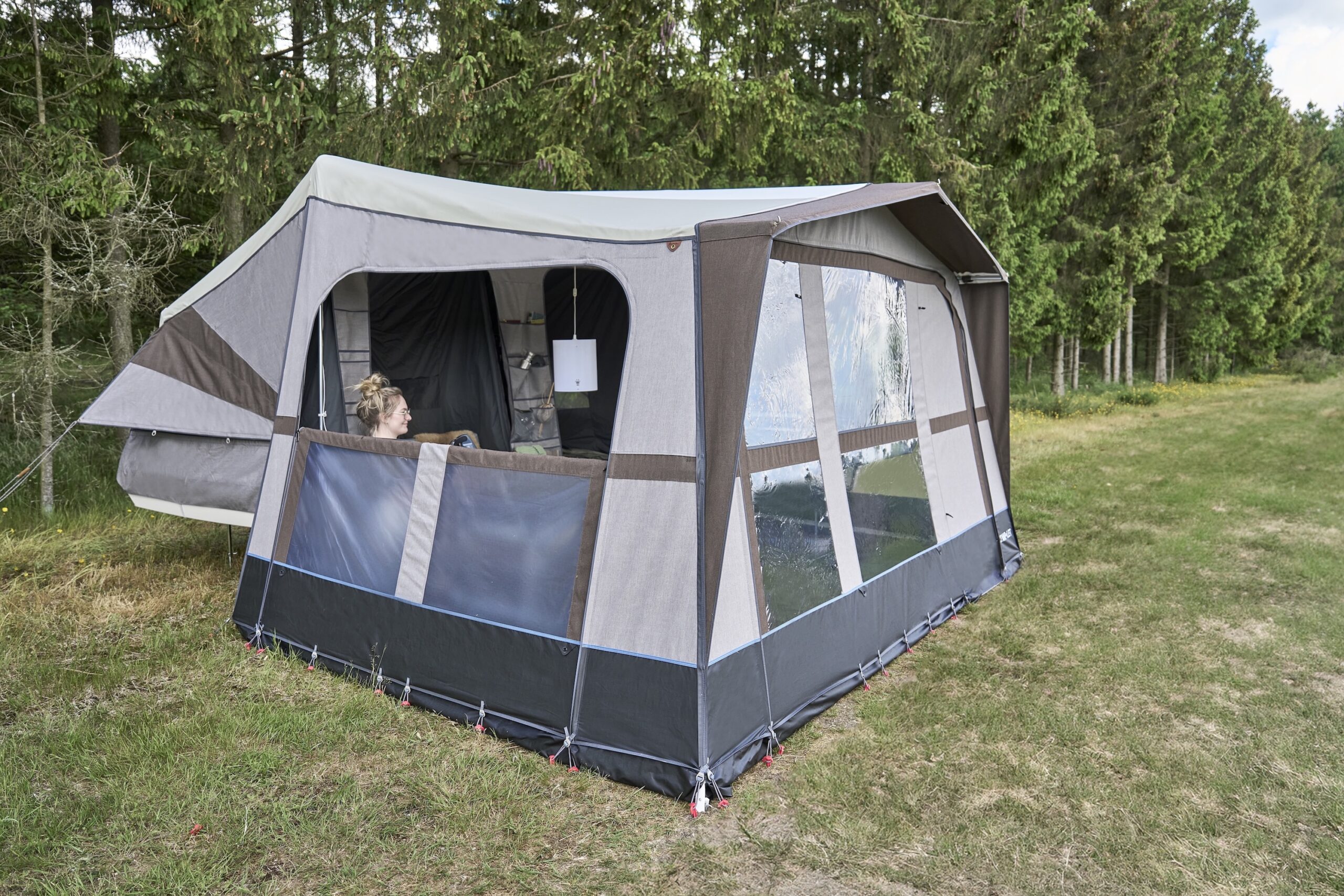 A spacious Camp-let tent is pitched on grass near trees, with a person sitting inside by an open window panel, showing the interior living space.