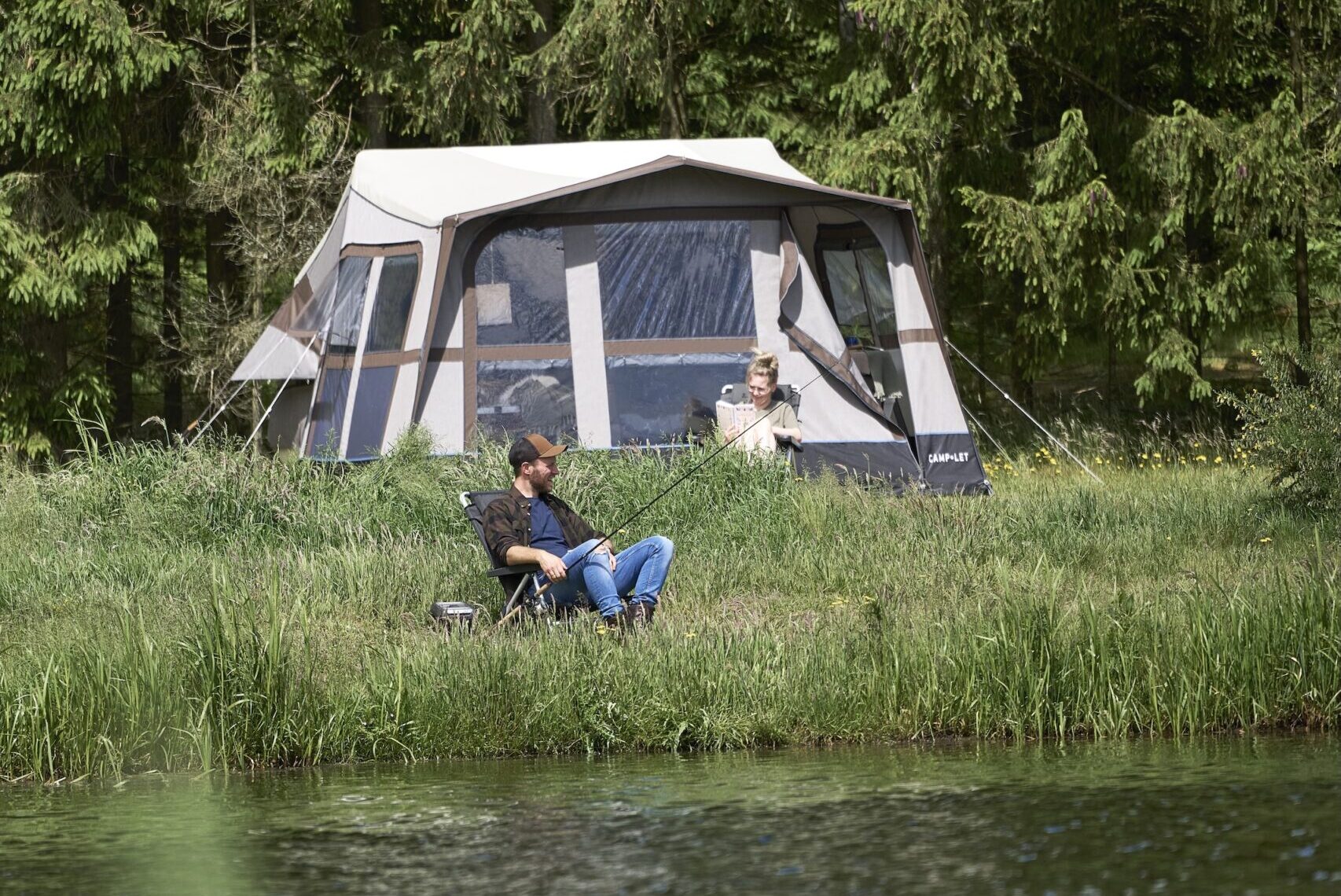 A Camp-let tent is fully set up beside a pond, with one person relaxing inside while another sits in a chair fishing at the water’s edge.