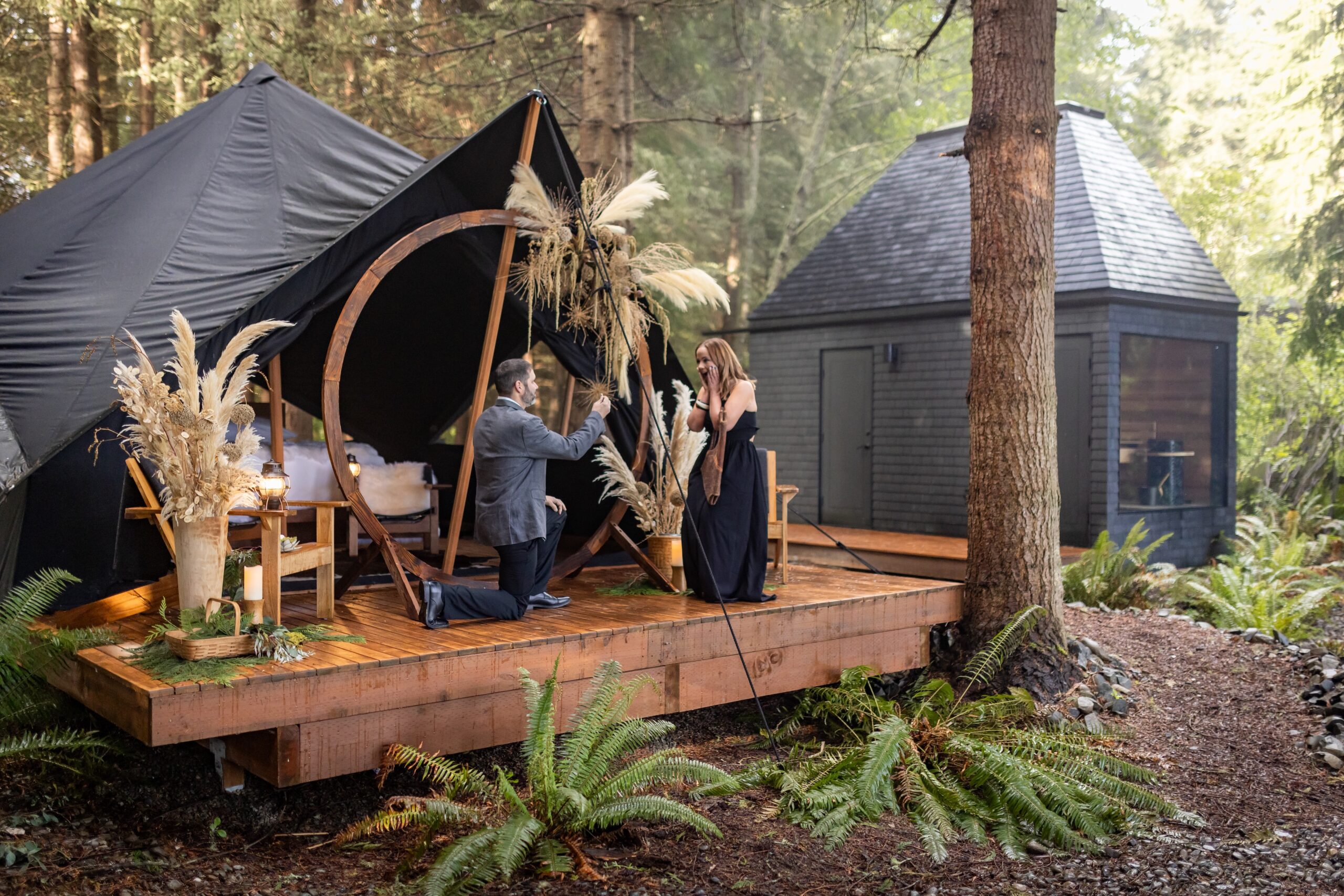 Romantic proposal at the Menizei luxury forest glamping retreat, with a man kneeling before a woman on a wooden deck beside a dark tent and floral arch.